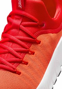 Rode sportieve schoen met een gestructureerde bovenkant van stof, voorzien van contrasterende oranje accenten, platte veters en een witte tussenzool.