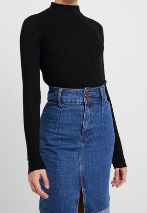 Femme portant un pull noir côtelé à col roulé et manches longues, rentré dans une jupe en jean bleue taille haute avec une fente devant et une fermeture à double bouton.