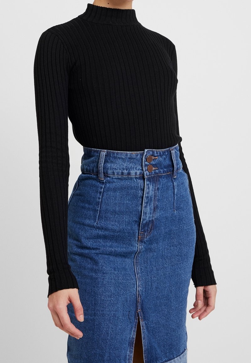 Femme portant un pull noir côtelé à col roulé et manches longues, rentré dans une jupe en jean bleue taille haute avec une fente devant et une fermeture à double bouton.
