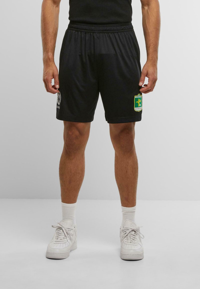 Man wearing black sports shorts with team logos, white socks, and white sneakers, standing against a plain light background.