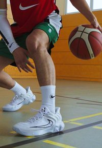 Valged korvpallijalatsid mustade aktsentidega ja tekstuuriga tallaga, kantud valgete sokkidega ja roheliste spordilühikesed püksidega, driblides korvpalli.