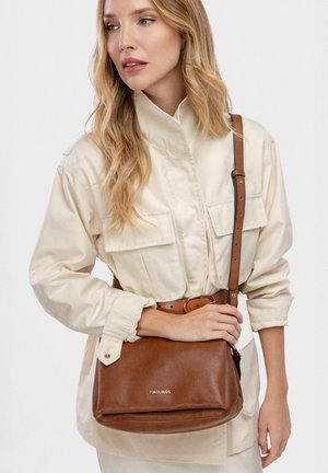 Femme blonde en veste beige tenant un sac bandoulière en cuir marron avec un logo "Pikolinos" visible.