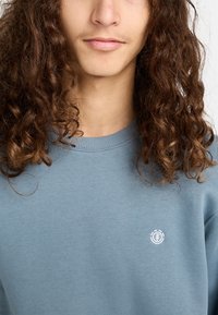 Sudadera azul grisácea con cuello redondo, hecha de una tela de textura suave. Presenta un pequeño logo blanco en la zona del pecho izquierdo.