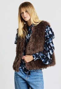 Kunstpelzweste in Braun mit flauschiger Textur und offenem Frontdesign. Getragen über einer blauen, gemusterten Bluse mit Rüschenärmeln.