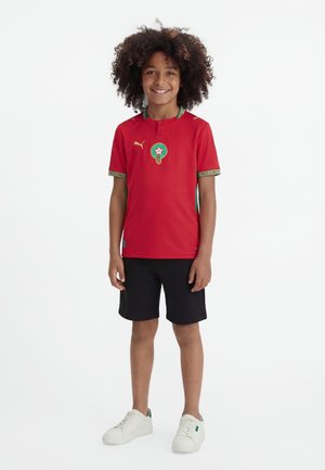 Χαμογελαστό παιδί με κατσαρά μαλλιά, φορώντας κόκκινη φανέλα ποδοσφαίρου, μαύρο σορτς και λευκά αθλητικά, στέκεται μπροστά σε ένα απλό λευκό φόντο.