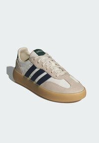 Béžová a bílá teniska s námořnicky modrými pruhy, z materiálů semiš a kůže, se zaoblenou špičkou a gumovou podrážkou. Na jazyku je zelené logo.