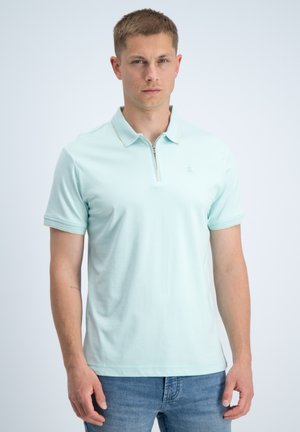 Junger Mann mit kurzen Haaren, der ein hellblaues kurzärmeliges Polohemd mit Reißverschluss und blaue Jeans trägt, steht vor einem schlichten Hintergrund.
