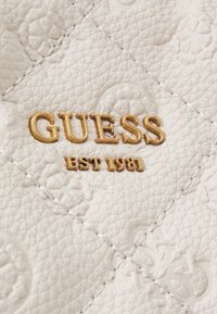 Cuir blanc texturé avec motifs floraux en relief et lettres métalliques dorées formant GUESS EST 1981 cousues dans la matière.
