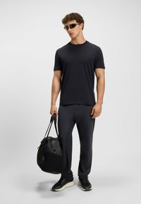 Schwarzes Kurzarm-T-Shirt und passende schmal geschnittene Hose, kombiniert mit schwarzen Sneakern. Trägt eine schwarze Sporttasche und trägt dunkle Sonnenbrillen.