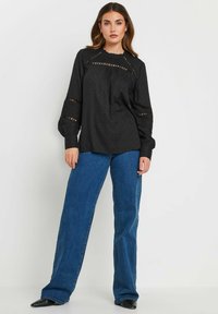 Blusa nera in tessuto testurizzato, con dettagli a taglio e maniche a sbuffo, abbinata a jeans in denim blu a vita alta e scarpe nere.