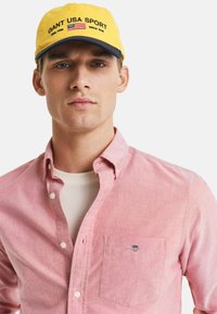 Gelbe Kappe mit "GANT USA SPORT"-Logo und amerikanischer Flagge; rosa Hemd mit Knöpfen, Tasche und subtiler Textur; weißes T-Shirt darunter.