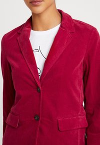 Nainen, jolla on päällään syvän vaaleanpunainen corduroy-puku takki valkoisen paidan päällä, jossa on minimalistinen musta kasvoviivapiirros.