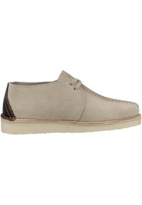 Beige mockaskinnssko med brunt läderackent vid hälen, snörningsdesign och beige gummisula. Har dekorativ sömnad längs sömmarna.