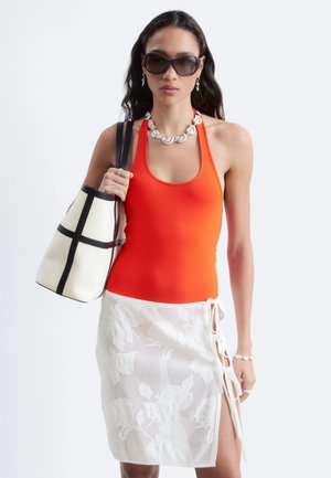 Femme portant de grandes lunettes de soleil, un top à col halter orange et une jupe portefeuille en dentelle blanche, tenant un sac blanc à rayures noires sur son épaule.