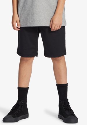 Shorts en coton noir avec une coupe droite, associés à des baskets montantes noires et des chaussettes côtelées noires, sous une chemise grise à manches courtes.
