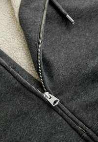 Graue Zip-Hoodie mit weichem cremefarbenem Fleece-Innenfutter. Verfügt über einen Metallreißverschluss und eine strukturierte äußere Stoffoberfläche. Nahaufnahme, die die Nähdetails zeigt.