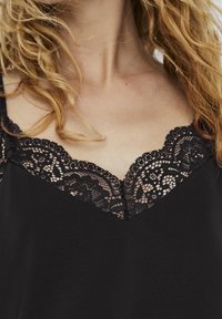 Svart linne med V-ringning i spets, som har intrikata blommönster. Materialet verkar slätt och något glansigt, med delikata spetsdetaljer.