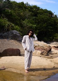 Femme en costume blanc et lunettes de soleil, debout pieds nus sur une plage de sable avec une côte rocheuse et une forêt verte en arrière-plan sous un ciel bleu.