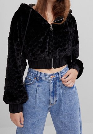 Woman wearing cropped black plush zip-up hoodie and high-waisted blue jeans, standing against plain light background.