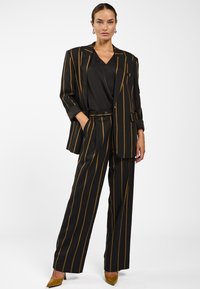 Zwart en gouden gestreepte pak met een losvallende blazer met een enkele knoopsluiting en brede broekspijpen. Soepele, op maat gemaakte stoftextuur.