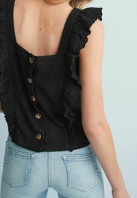 Sort stoftop med flæse-blondekant, åben ryg og knaplukning. Kombineret med lyseblå tætsiddende jeans.