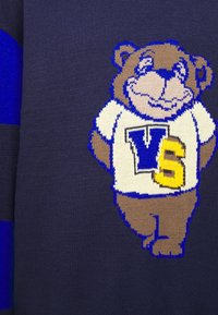 Pull bleu marine avec un ours tricoté beige, portant une chemise blanche à bordure bleue, affichant les lettres "VS" en bleu et jaune.