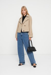 Veste beige cropped avec un large col et deux boutons foncés, portée sur un haut noir. Jean bleu taille haute et chaussures plates noires. Sac à main noir.
