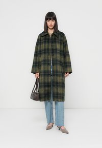 Grønn og svart rutete ullfrakk med krage, frontknapper og sidelommer; brukt over lyseblå utsvingte jeans og beige hæler.