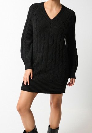 Femme portant une robe-pull noire en maille torsadée à col en V avec manches longues et des bottines noires, sur fond blanc.