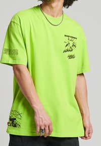 Neonově zelené bavlněné tričko s krátkými rukávy, s potiskem na přední části a designem na zadní straně, zobrazujícím ostnatý drát a postavu.