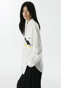 Chemise blanche texturée avec une fermeture à boutons, ornée d'une broderie d'un chat noir et d'un citron jaune sur la poche gauche de la poitrine. Manches longues.