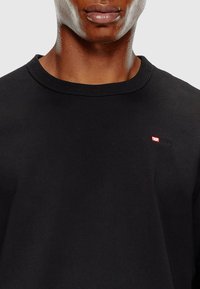T-shirt en coton noir avec un col rond, comportant un petit logo Diesel rouge et blanc sur la poitrine gauche. Texture lisse, coupe décontractée.