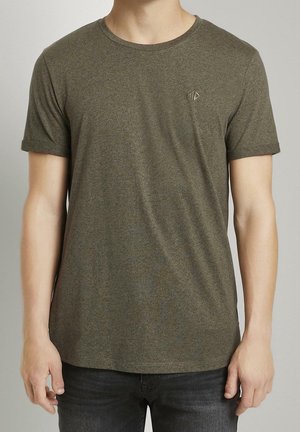Homme portant un t-shirt uni vert olive à manches courtes avec un petit logo sur la poitrine gauche, associé à un jean foncé.
