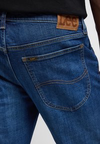 Temno modri jeans iz denima z rjavim usnjenim oznako "Lee" na pasu, z vidnimi šivi in detajli na zadnjem žepu.