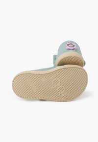 Scarpa in tessuto azzurro chiaro con suola in gomma beige morbida, dotata di un'etichetta rettangolare sul retro con un design di cartone animato e testo.