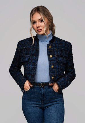 Chaqueta de tweed azul marino con un acabado texturizado, botones dorados y bolsillos frontales, combinada con un jersey de cuello alto de color azul claro y vaqueros de cintura alta.