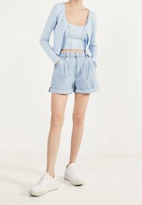 Person klädd i en ljusblå, kort cardigan över en matchande tanktop, högmidjade, nerullade denimshorts och vita sneakers, står med händerna i fickorna.