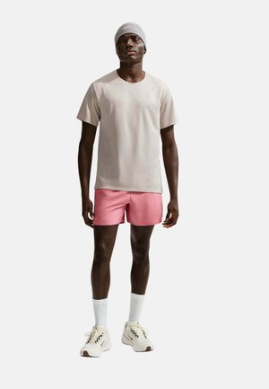 Hombre con camisa beige claro de manga corta, pantalones cortos rosas, calcetines blancos, zapatillas deportivas y un gorro gris de Nike, de pie sobre un fondo blanco.