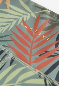 Tessuto a fantasia con foglie tropicali verdi, ruggine e blu scuro con la scritta "ACTIVE" in oro su uno sfondo blu tenue.