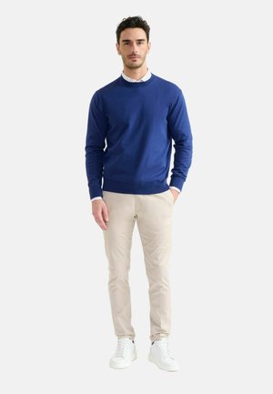 Uomo in piedi che indossa un maglione blu sopra una camicia bianca, pantaloni beige e scarpe da ginnastica bianche su sfondo chiaro uniforme.