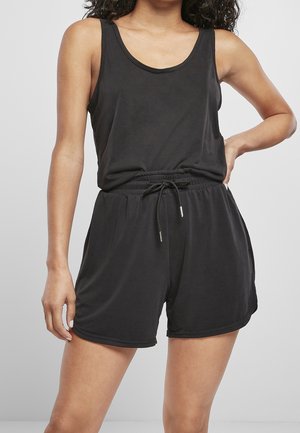 Kobieta ubrana w czarny, bezrękawnik i czarne szorty z wiązanym pasem, stoi z jedną ręką na biodrze na jednolitym tle.