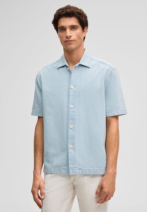 Mężczyzna z kręconymi brązowymi włosami, ubrany w jasnoniebieską koszulę na krótki rękaw z guzikami i białe spodnie, stojący na jednolitym tle.