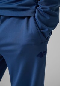 Gros plan d'une personne portant un pantalon de sport bleu avec les mains dans les poches latérales et un logo "4F" visible sur la cuisse.