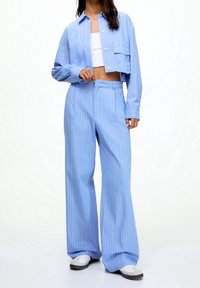 Kobieta ubrana w jasnoniebieską marynarkę w prążki o skróconym fasonie oraz dopasowane szerokie spodnie, do tego białe sneakersy i biały top. 