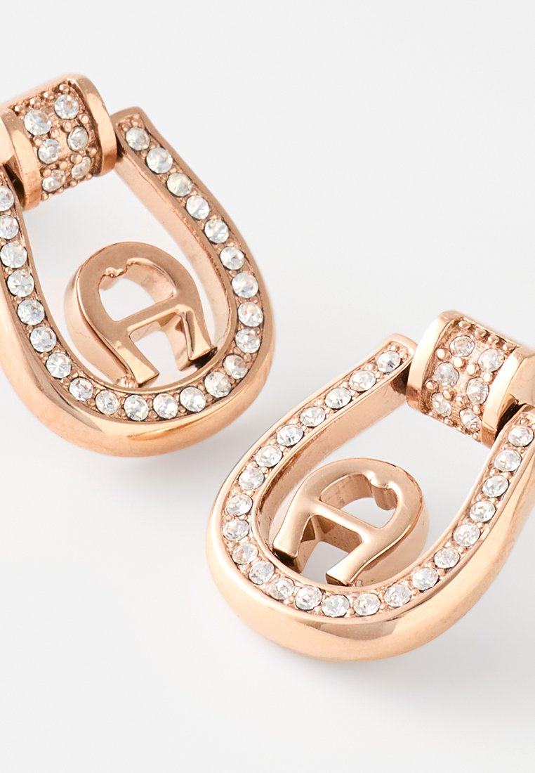 Ohrringe in Roségold in Form eines Hufeisens, verziert mit klaren Strasssteinen und mit einem stilisierten "A" in der Mitte. Glatte Oberfläche.