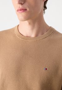 Maglione lavorato a maglia marrone chiaro con una superficie testurizzata, scollo rotondo e un piccolo logo multicolore sul petto.