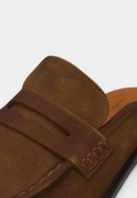 Κοντινό πλάνο ενός καφέ δερμάτινου μοκασίνι με λουράκι για κέρματα και λεπτομερή ραφές σε λευκό φόντο.