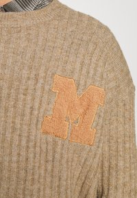 Béžový pletený svetr s žebrovanou strukturou. Na levé straně má chlupatou oranžovou nášivku ve tvaru "M". Kulatý výstřih a měkký materiál.