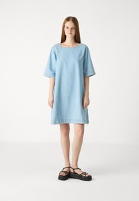 Robe en denim bleu clair avec une coupe ample, des manches courtes, un col rond et un ourlet effiloché. Associée à des sandales noires à semelle épaisse.