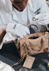 Personne portant des gants blancs et une blouse de laboratoire examine le contenu d'un sac à main beige à l'aide d'une loupe et d'un outil.